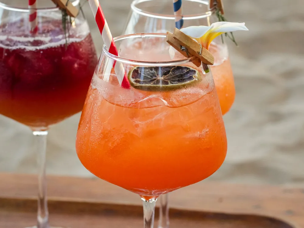 To farverige cocktails med sugerør og frugtpynt i nærbillede, med en blød strand i baggrunden