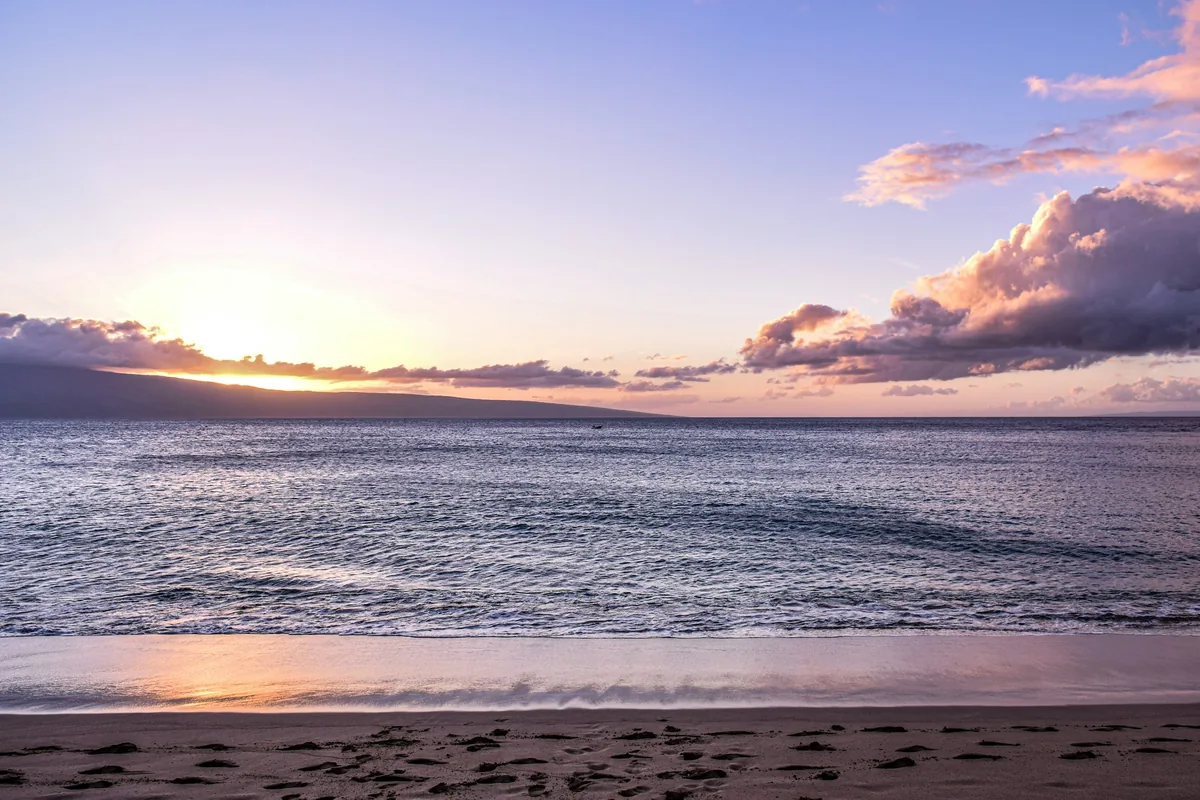 Solen står lavt over et stille hav med den mørke silhuet af West Maui og bløde violette skyer derover
